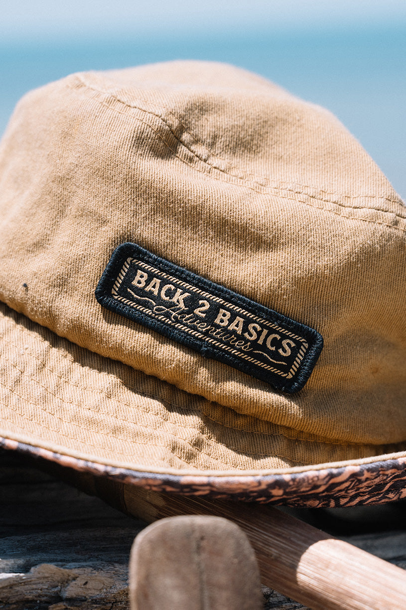 Beige bucket hat with 'Back 2 Basics' logo against a blurred natural background