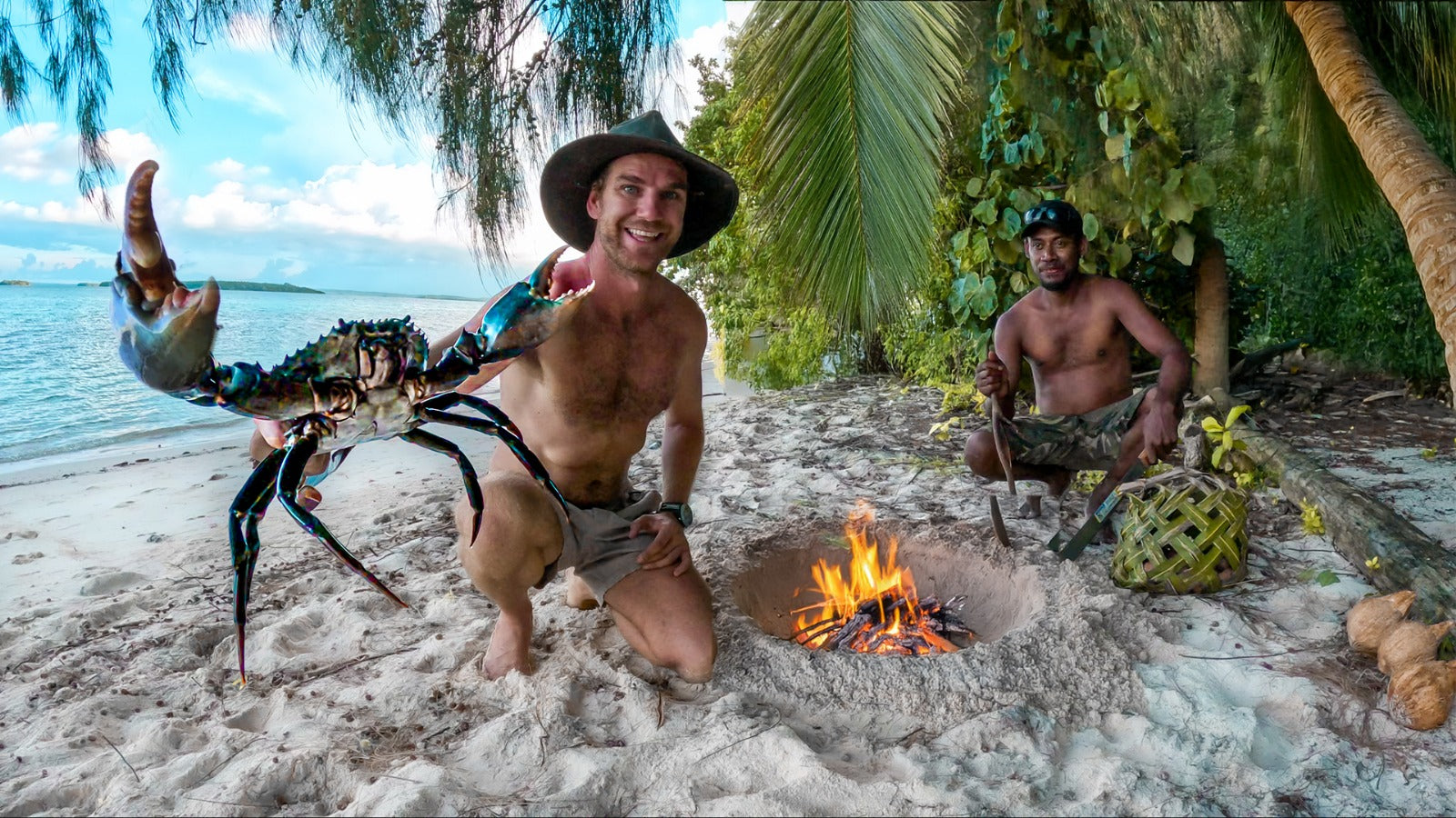 Two men on a beach with a large crab and a fire pit, surrounded by tropical vegetation.