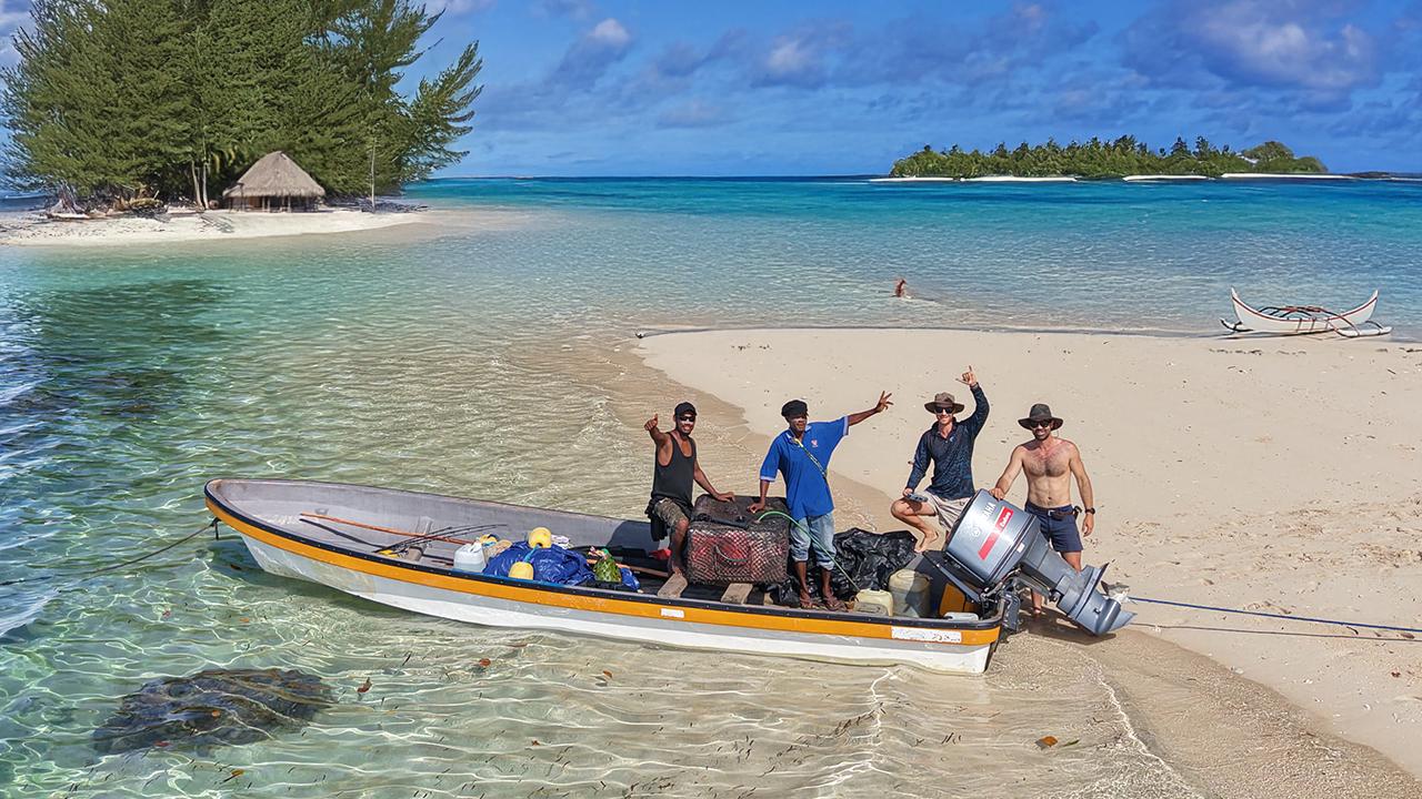 People with a boat on a beach with clear water and a tropical island in the background