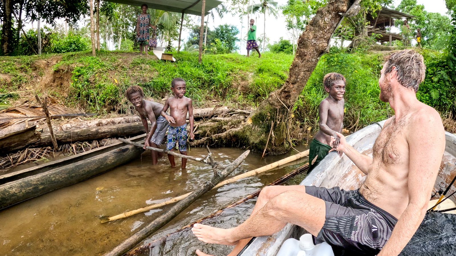 Man sitting on a log in a river with children playing nearby, surrounded by greenery.