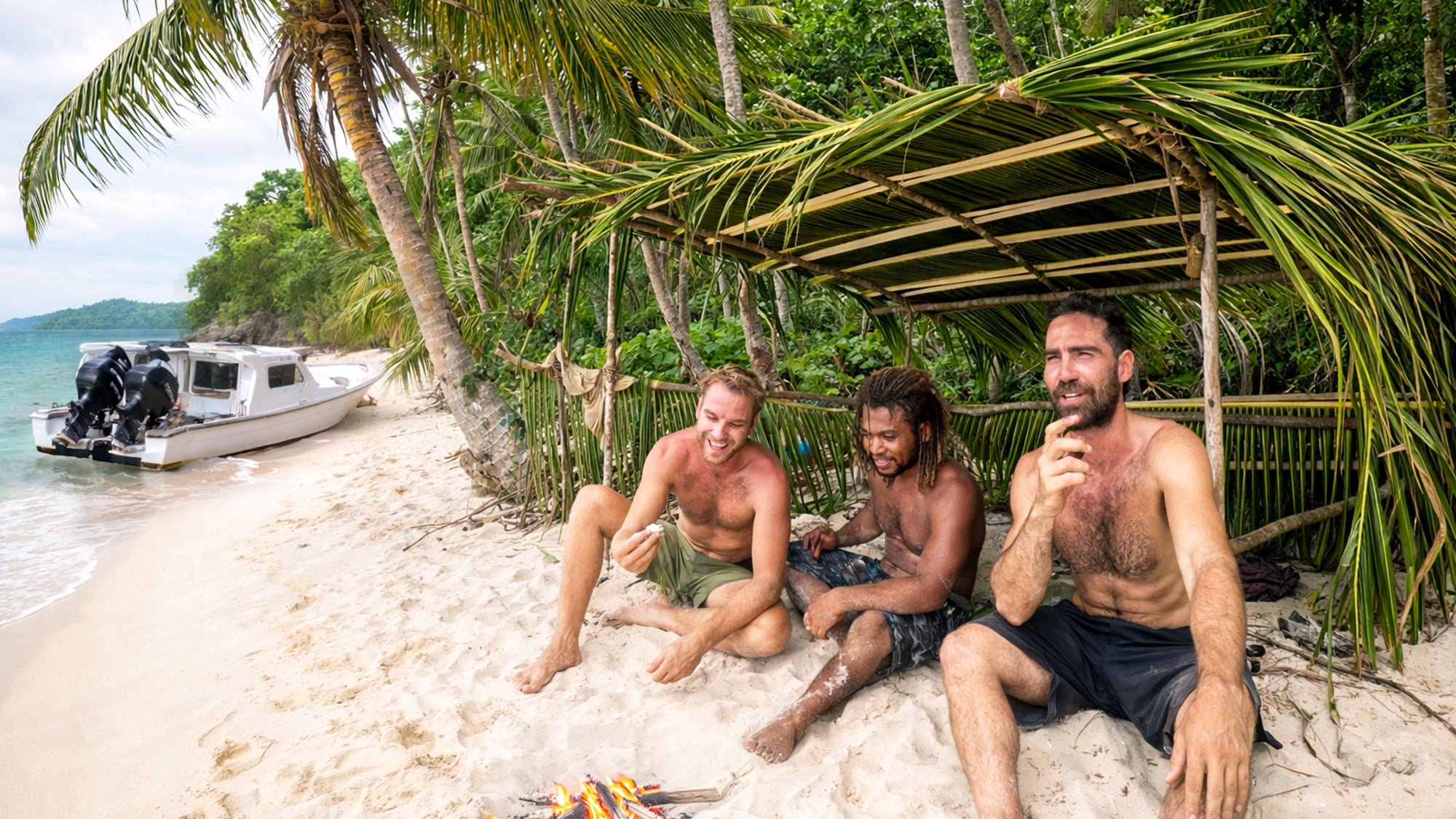 Three men sitting by a campfire on a tropical beach with a boat and palm trees in the background.