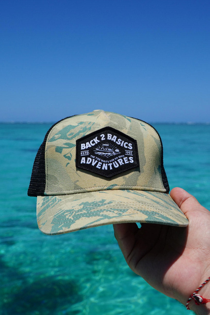 Camouflage cap with 'Back 2 Basics Adventures' patch held against a clear blue ocean background