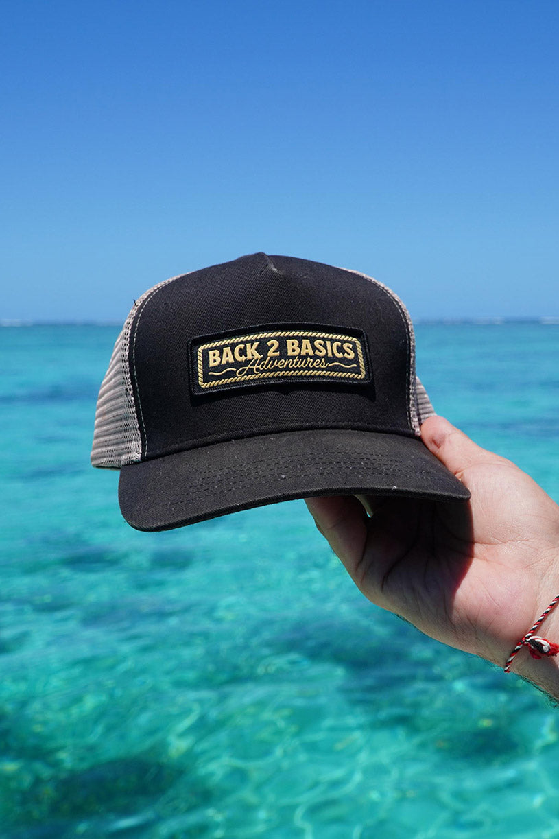 Black cap with 'Back 2 Basics' logo held against a clear blue ocean background