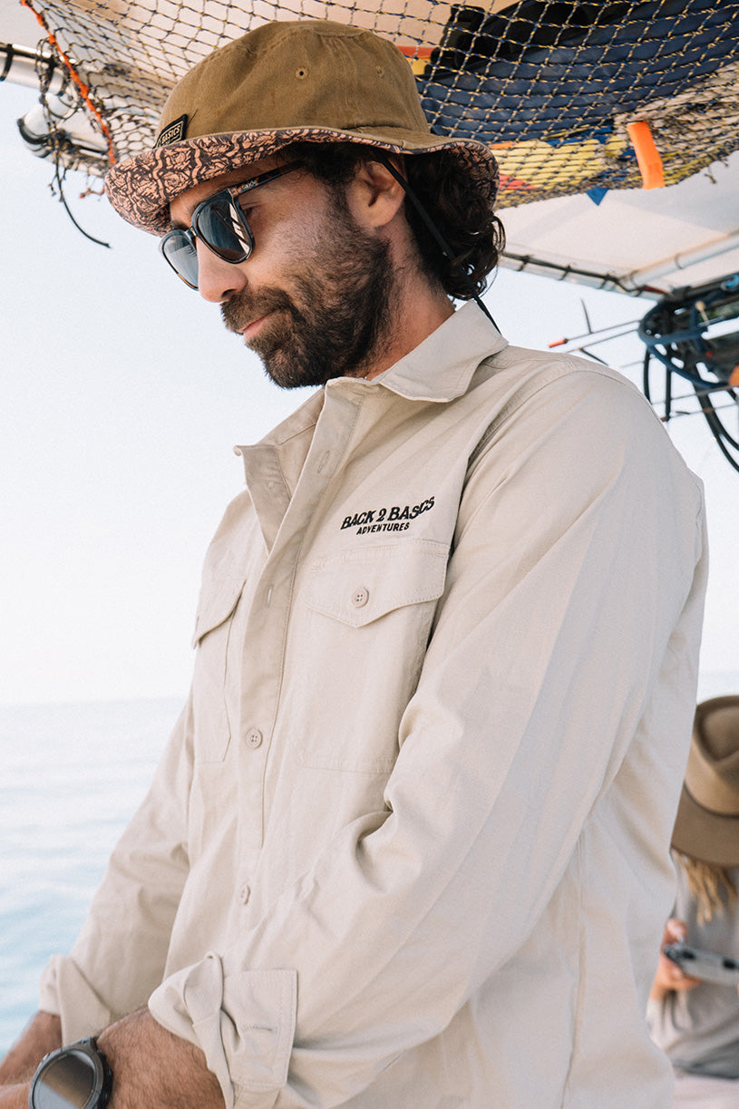 Man wearing a beige fishing shirt with a b2b adventures logo, sunglasses, and a hat on a boat.