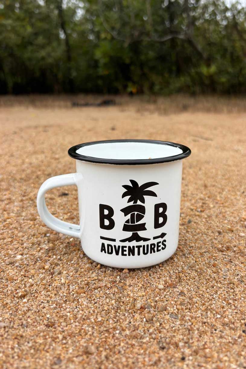 White mug with 'B&B Adventures' logo on sandy ground with greenery in the background