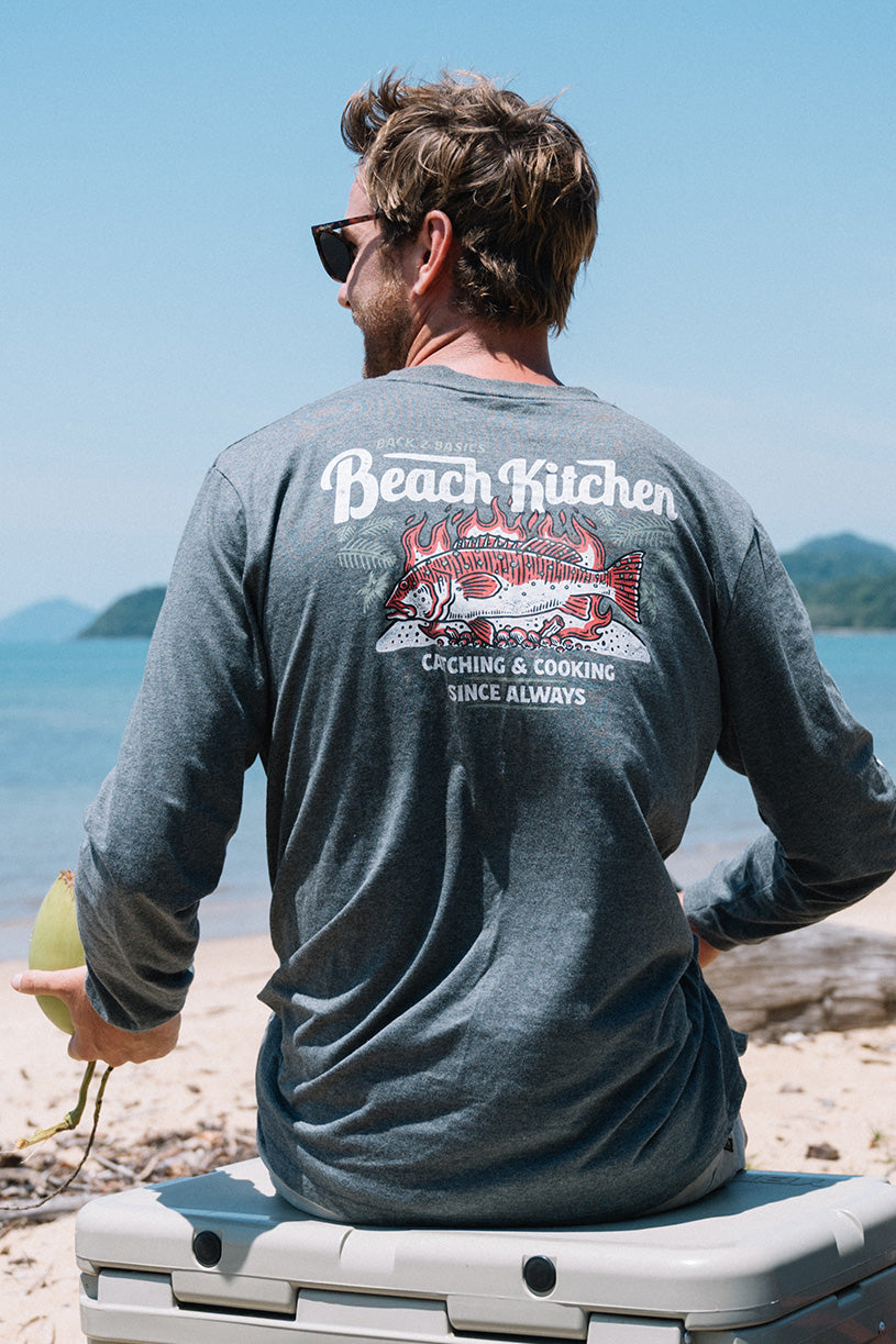 Man wearing a 'Beach Kitchen' shirt sitting on a cooler by the beach.